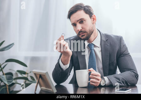 Trauriger Mann im grauen Anzug an den hölzernen Tisch sitzen mit Smartphone, Foto im Rahmen halten weiße Kappe und Ring in Zimmer, trauernde Unordnung Konzept Stockfoto