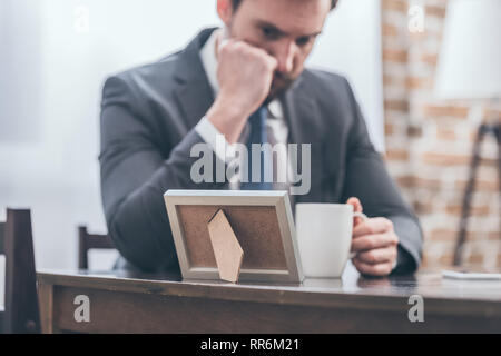 Selektiver Fokus der umgekippt Mann im grauen Anzug an den hölzernen Tisch sitzen, halten weiße Tasse und Bilderrahmen in Zimmer, trauernde Unordnung Konzept Stockfoto