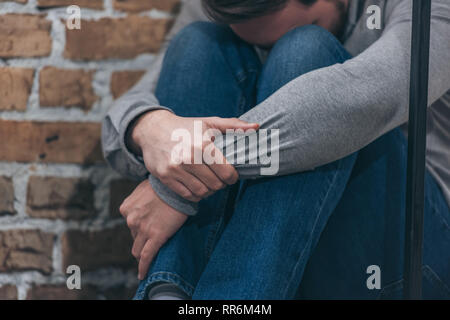 7/8-Ansicht der Mann im grauen Pullover und blauen Hosen sitzen und umarmen Knien auf Braun strukturierten Hintergrund in Zimmer, trauernde Unordnung Konzept Stockfoto