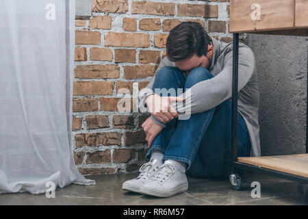 Verärgert Mann sitzt auf dem Boden in der Ecke und umarmen Knie mit den Kopf auf Braun strukturierten Hintergrund in Zimmer, trauernde Unordnung Konzept Stockfoto