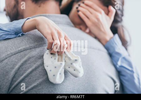 7/8-Ansicht von Frau, umarmen, Mann, Holding Baby Socken und Weinen in Zimmer, trauernde Unordnung Konzept Stockfoto