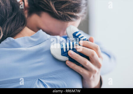 7/8-Ansicht von Mann, Frau, Umarmen und Halten blau Baby Schuhe im Zimmer, trauernde Unordnung Konzept Stockfoto