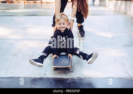 Junge Mutter lehrt, ihren kleinen Jungen ein Skateboard zu fahren Stockfoto