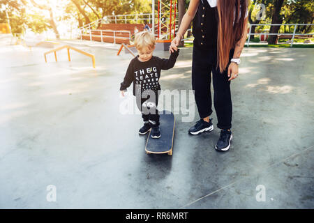 Junge Mutter lehrt, ihren kleinen Jungen ein Skateboard zu fahren Stockfoto