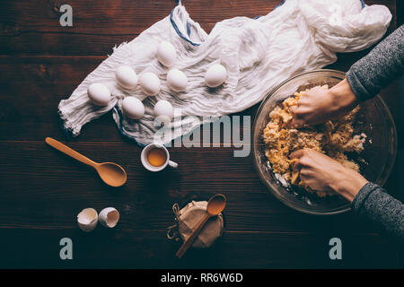 Woman's Hand kneten den Teig in der Schüssel Neben Eiern, Löffel und marmeladenglas. Blick von oben auf braun Küche aus Holz Tisch mit Zutaten zum Kochen pie Stockfoto