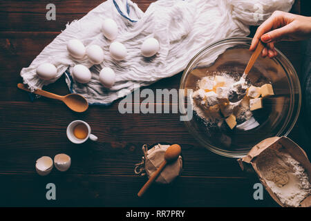 Blick von oben auf braun Küche aus Holz Tisch mit Zutaten zum Kochen pie. Woman's Hand streusel Mehl auf gewürfelte Butter in Schüssel Neben Eiern, spo Stockfoto
