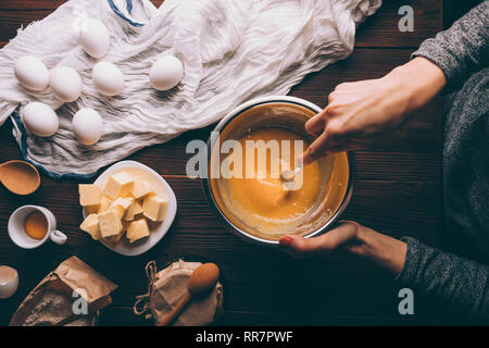 Die Ansicht von Oben weibliche Hände rühren, Zucker und Eigelb in Metall Schüssel weiter Butter, Mehl und Eiern auf braunem Holz- Küche Tabelle gewürfelt. Prozess des Kochens Stockfoto