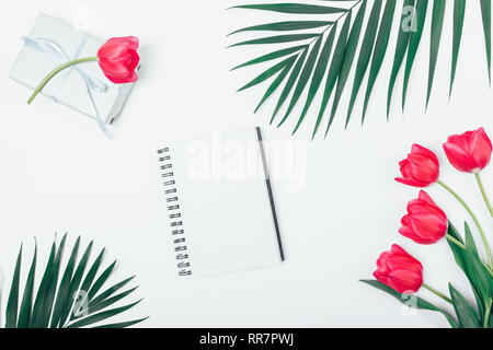 Flach Anordnung von leer Notebook unter Palmen Blätter und Blüten rosa mit Geschenkbox auf weißem Tabelle, Ansicht von oben. Stockfoto