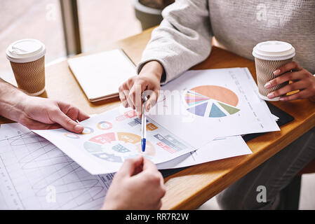Nahaufnahme von zwei Kollegen ihre Arbeit diskutieren über Holzschreibtisch. Sie studieren die Charts. Frau hält einen Stift und eine Tasse Kaffee. Lässigen Stil. Konzept der Erfolg Stockfoto