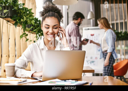 Gerne afrikanische amerikanische Frau sitzt an der hölzernen Büro Schreibtisch, am Laptop arbeiten, in angenehmen Gespräch oder Zelle Gespräch. Attraktives Mädchen lächelt, während sie ein Smartphone am Arbeitsplatz. Stockfoto