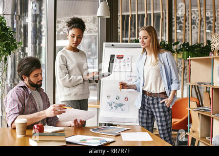 Attraktive junge Frau, die in der Nähe der Tafel mit einem Laptop in der Hand und Suchen zu ihren männlichen Kollegen, die ein Diagramm mit den Ergebnissen. Ein anderer Kollege schaut auf sie mit einem Lächeln. Konzept der Erfolg Stockfoto