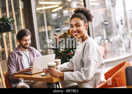 Zwei freundliche Mitarbeiter sitzen über Schreibtisch. Bärtige kaukasischen Mann mit seinem Laptop und attraktive afrikanische amerikanische Frau lächelnd in die Kamera, Kaffee halten und schreiben neue Ideen. Lässigen Stil. Konzept der Erfolg Stockfoto