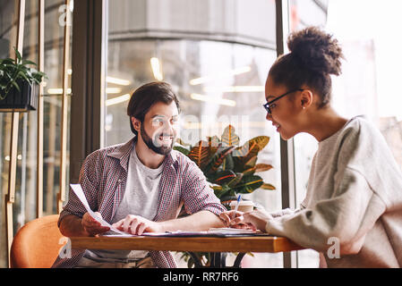 Zwei freundliche Kollegen diskutieren neue Ideen über Schreibtisch sitzen. Bärtige kaukasischen Mann und attraktive afrikanische amerikanische Frau mit Diagrammen arbeiten. Kaffee, Arbeitspapiere und Bleistifte werden auf dem Schreibtisch. Lässigen Stil. Konzept der Erfolg Stockfoto
