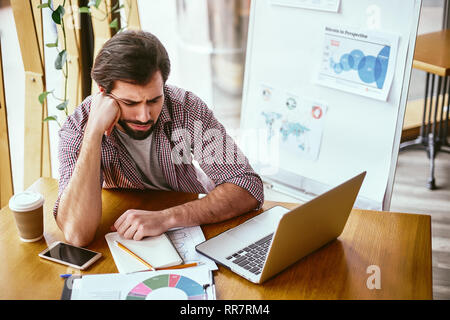 Bärtiger junger Mann in lockerer sitzt an seinem Arbeitsplatz und Ruhen, den Kopf auf den Arm. Porträt einer müde Junge Unternehmer am Tisch sitzen mit Laptop in der Verzweiflung. Modernes Büro Stockfoto