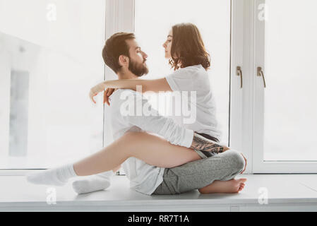 Schöne junge Paare an der jeweils anderen suchen, umarmen und sitzen auf Fensterbank zu Hause Stockfoto