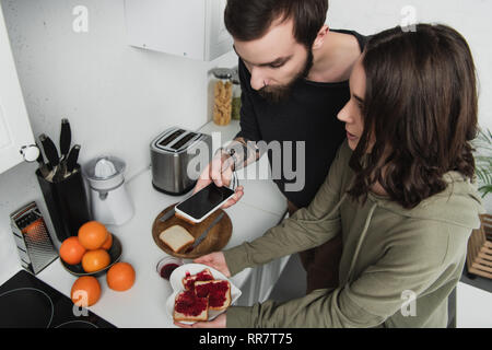 Schöne junge Paar unter Foto von Toast auf dem Smartphone beim Frühstück in der Küche Stockfoto