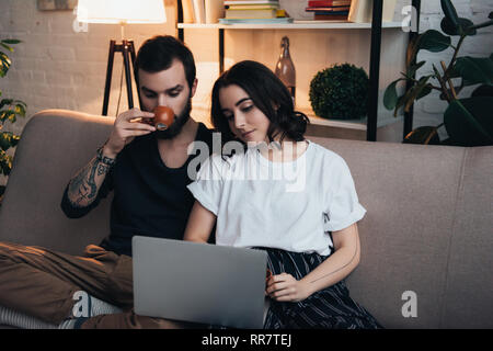 Mann sitzt auf der Couch und tranken Kaffee, während schöne Frau mit Laptop im Wohnzimmer Stockfoto