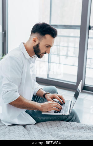 Schöner mann Tippen auf Laptop Tastatur im Schlafzimmer konzentriert Stockfoto