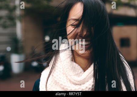 Lächelnde Frau auf der Straße mit Haare fliegen im Gesicht. Nahaufnahme einer asiatischen Frau in Winterkleidung stehen draußen mit ihr Haar bedecken Ihre f Stockfoto