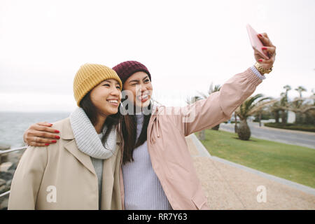 Zwei asiatische Frauen im Winter Kleidung am Meer stehen und eine selfie mit Handy. Frauen Freunde Spaß im Freien an einem kalten Tag. Stockfoto