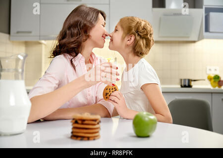 Glückliche Mutter und Tochter auf Küche zusammen sitzen, ein Lächeln auf den Lippen. Schöne Frau mit Milch Glas, kleine Mädchen Holding american Cookie. Kekse mit Stockfoto