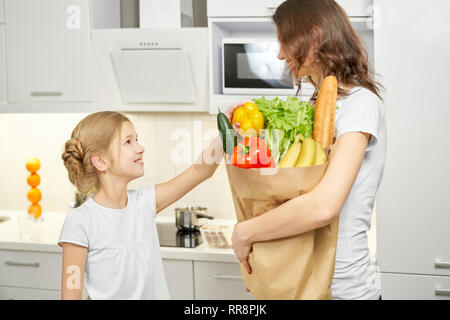 Frau mit Papiertüte mit frischen Produkten nach dem Einkaufen. Nette, hübsche Tochter stehend mit Mama in Weiß, moderne Küche. Gemüse, Grüns, Pepp Stockfoto