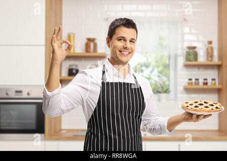 Lächelnden jungen Mann mit einer Schürze Holding eine Torte in einer Küche und deutete mit der Hand Stockfoto