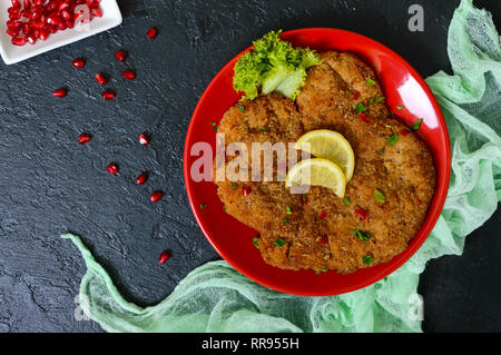 Große Wiener Schnitzel auf einer roten Platte mit Zitrone auf einem schwarzen Hintergrund. Fleischgericht. Ansicht von oben, flach. Stockfoto