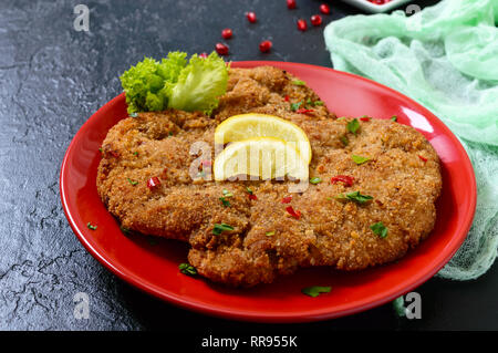 Große Wiener Schnitzel auf einer roten Platte mit Zitrone auf einem schwarzen Hintergrund. Fleischgericht. Stockfoto