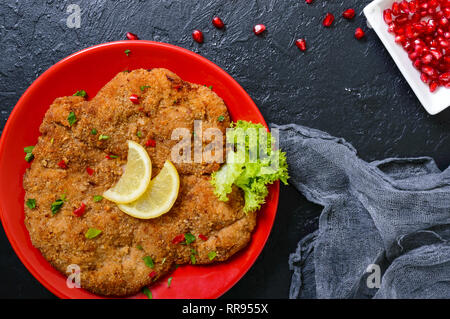 Große Wiener Schnitzel auf einer roten Platte mit Zitrone auf einem schwarzen Hintergrund. Fleischgericht. Ansicht von oben, flach. Stockfoto