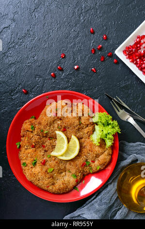 Große Wiener Schnitzel auf einer roten Platte mit Zitrone auf einem schwarzen Hintergrund. Fleischgericht. Ansicht von oben, flach. Stockfoto