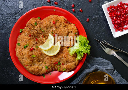 Große Wiener Schnitzel auf einer roten Platte mit Zitrone auf einem schwarzen Hintergrund. Fleischgericht. Ansicht von oben, flach. Stockfoto