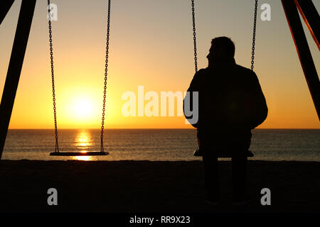 Rückansicht Hintergrundbeleuchtung Silhouette, ein Mann, der allein auf einer Schaukel bei leerem Sitz bei Sonnenuntergang am Strand im Winter Stockfoto
