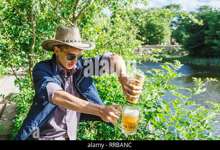 Junger Mann in einen Cowboy Hut und Sonnenbrille ist Bier zapfen aus einem jar in einem Becher im Park auf sonnigen Sommertag Stockfoto