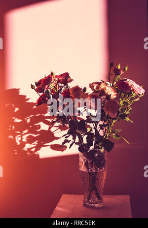 Ein Blumenstrauß aus Rosen in einer Glasvase stehen auf einer hölzernen Schemel vor dem Hintergrund der Wand in den Strahlen des Sonnenlichts Stockfoto