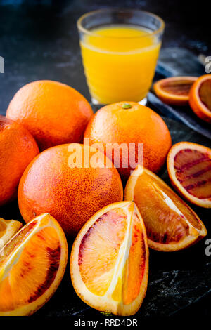 Frischen saftigen Blutorangen Ganze und Aufgeschnittene auf dunklen Platte mit Glas Orangensaft und Schichten im Hintergrund - geringe Tiefenschärfe Stockfoto