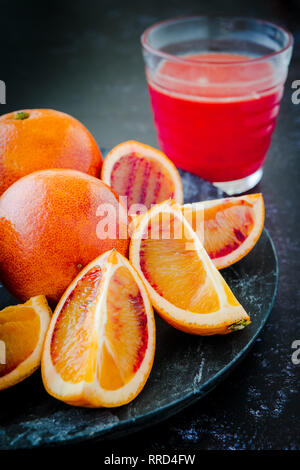 Frischen saftigen Blutorangen Ganze und Aufgeschnittene auf dunklen Platte mit Glas Saft im Hintergrund - geringe Tiefenschärfe Stockfoto