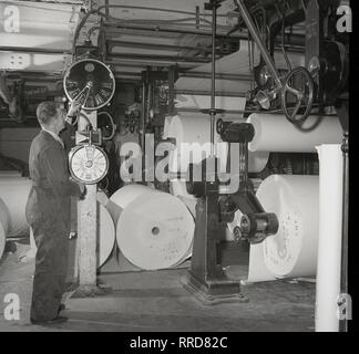 1950er Jahre, männlichen Arbeitnehmer in Latzhosen Kontrolle der Zifferblätter und Bedienelemente in einem Print funktioniert Der brittains Papierfabrik, England, UK, in dem die Maschinen, bei denen die fertigen Papierrollen gelagert werden. Stockfoto