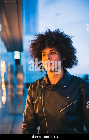Hübscher junger Mann mit afro Haar durch gelbe Licht eines Showcase ernsthaft mit dem städtischen Hintergrund in blau beleuchtet Stockfoto