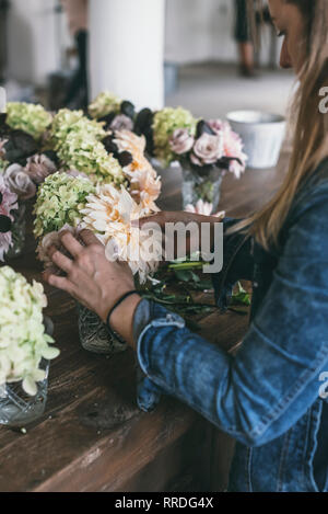 Glückliche Dame in der Nähe von Holz- Tabelle mit Büscheln von frischem Chrysanthemen, Rosen und Pflanze Zweige in Vasen auf grauem Hintergrund Stockfoto