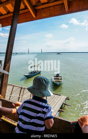 Blick auf unscharfen Vordergrund Kinder beobachten das Boot von den Holzsteg mit Johor River Bridge im Hintergrund angedockt. Blick von einem Holzsteg Stockfoto