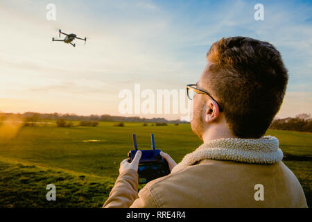 Ein männlicher drone Pilot mit einer Pilotenlizenz mit Hilfe einer Fernbedienung ein quadcopter Stil drone für Fotografie und Video Zwecke im Freien bei Sonnenuntergang zu fliegen Stockfoto