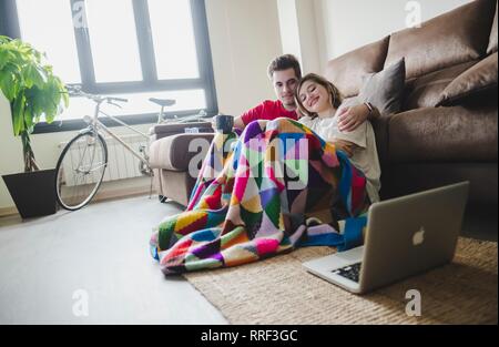Umarmen jungen Mann und Frau sitzen auf dem Boden auf der Couch und beobachten Laptop zu Hause Stockfoto