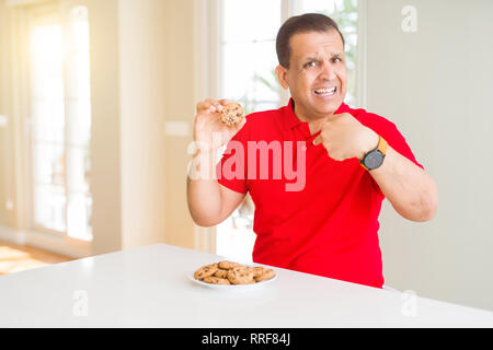 Mittleres Alter Mann essen Chocolate Chips cookies zu Hause mit Überraschung Gesicht zeigenden Finger zu sich selbst Stockfoto