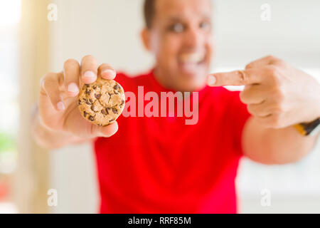 Mittleres Alter Mann essen Chocolate Chips cookies zu Hause sehr zufrieden zeigte mit Hand und Finger Stockfoto