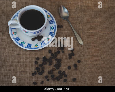 Frühstück eines Porzellan Kaffeetasse, einen antiken Löffel und Kaffeebohnen auf einem vintage Sackleinen Hintergrund Stockfoto