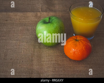 Frühstück aus einem Glas mit Orangensaft eine Mandarine und ein grüner Apfel auf einem vintage Sackleinen Hintergrund Stockfoto
