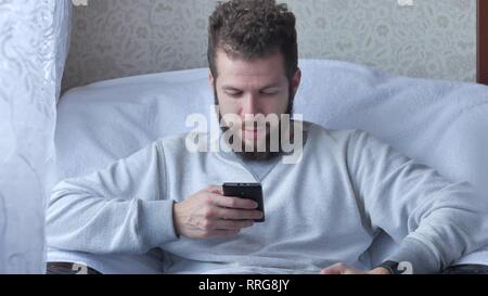 Ein weißer Junge brunette lockig-bärtigen Mann nutzt das Telefon in einem Stuhl sitzend Trinken von Tee oder Kaffee. Stockfoto