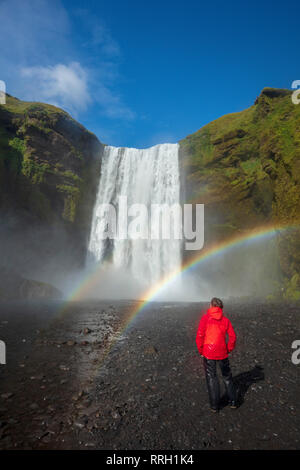 Rainbow und Person unter 60 m hohen Wasserfall Skogafoss. Skogar, Sudhurland, South Island. Stockfoto