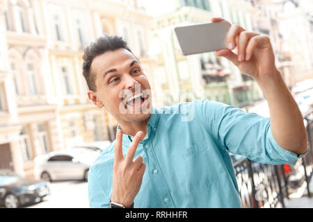 Erfassen Momente. Junger Mann auf der Straße der Stadt, die auf dem Smartphone angezeigt selfie Friedenszeichen in die Kamera lächeln schließen aufgeregt - Bis Stockfoto